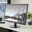 An AOC 2460SD 24 Certified Refurbished Monitor (1920x1080, HDMI, VGA, DVI, USB | NO STAND) displays a splash of water on a light-colored desk with blurred office items and a potted plant in the background.
