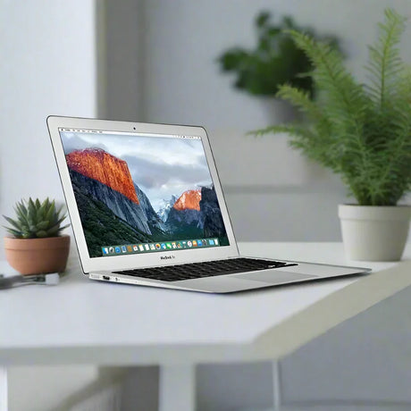 An Apple MacBook Air Early 2015 A1466 i5 5250U 1.6GHz 8GB 256GB SSD 13.3 (silver) sits open on a white desk, displaying a mountain scene, with a small cactus, a fern, and blurred green plants in the background.