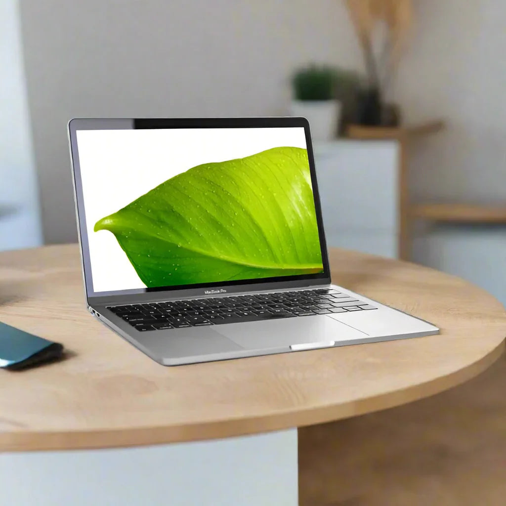 An Apple MacBook Pro Mid 2017 A1708 i5 7360U 2.3GHz 16GB 256GB SSD 13.3" | Wty sits on a round wooden table with its screen displaying a close-up of a green leaf, a smartphone nearby, and blurred furniture and plants in the background.