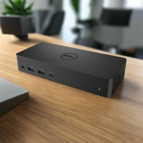 A certified refurbished Dell D6000 USB 3.0 HDMI Display Ports Docking Station by Dell rests on a wooden desk with visible front ports, office furniture and potted plants blurred behind—ideal for versatile multi-monitor setups. Adapter included.