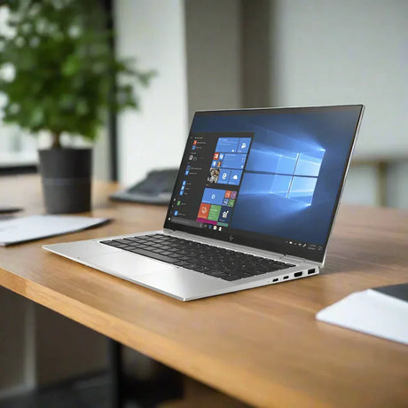 A silver HP EliteBook X360 1030 G7 i7 convertible laptop displays Windows 11 Pro on a wooden desk, with blurred office items and a plant in the background.