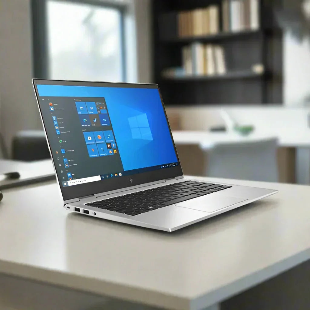 A silver HP EliteBook X360 830 G8 i5 1145G7 2.6GHz 8GB 256GB W11P 13.3" Touch laptop sits open on a white desk in a modern office, displaying Windows and softly blurred shelves and books in the background.