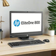 An HP EliteOne 800 G1 AIO i7 desktop with 8GB RAM, 1TB HDD, and a 23 display sits on a light wood desk with keyboard and mouse. The HP logo is on screen; plants and office supplies are in the background.