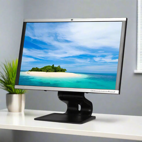 An HP LA2405W 24 1920x1200 monitor sits on a white desk, displaying a scenic island view. A green plant and a window with blinds are in the background. Brand: HP.