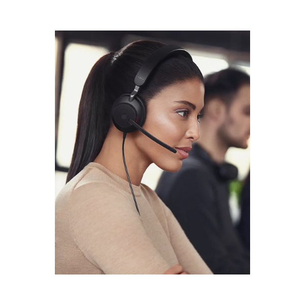 A woman with long dark hair in a ponytail, wearing a beige top and a Jabra Evolve2 40 Mono Wired Headset USB-C, sits focused in an office. Another person with a headset is visible in the background.