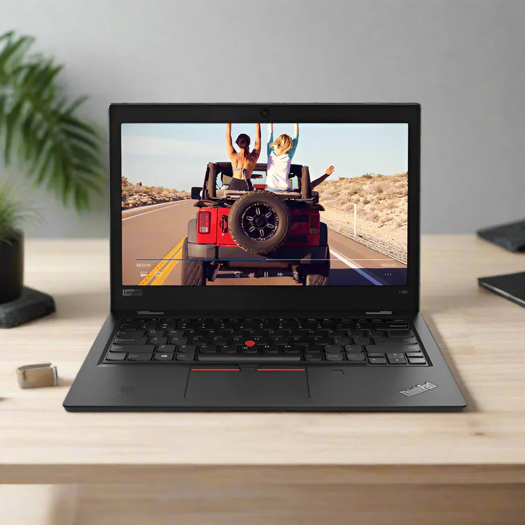A Lenovo ThinkPad L380 i5 8350U 13.3 sits on a wooden desk, displaying a photo of two people in a red Jeep on a sunny road with arms raised; its surrounded by a small plant and various desk items.