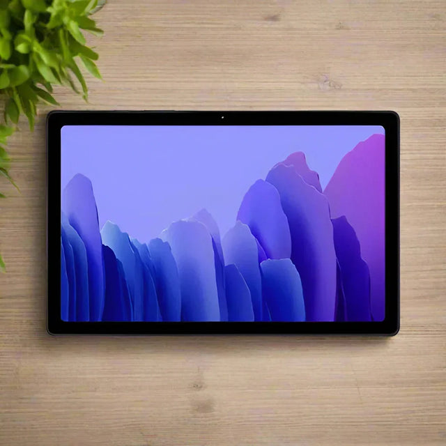 A certified refurbished Samsung Galaxy Tab A7 SM-T505 32GB Tablet Black by Samsung rests on a light wooden surface beside a green leafy plant, displaying a purple and blue abstract wallpaper.