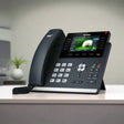 The Yealink SIP-T46S Gigabit IP Phone + Adapter | Wty by Yealink features a corded handset and color display, shown on a light desk in a blurred office setting, ideal for enhanced business communication.