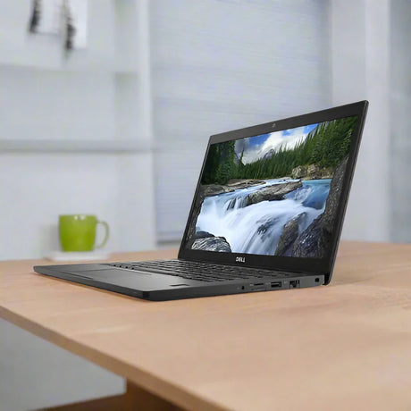 A Dell Latitude 7490 i5 8350U 1.7GHz 8GB 256GB SSD W11P 14 Laptop sits on a wooden table, showing a waterfall and forest. A green mug and shelves appear blurred in the background against a light-colored wall.