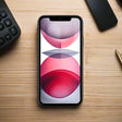 An Apple iPhone 11 64GB Black Unlocked Smartphone with a cracked back and battery sits on a light wooden desk with a red and gray abstract wallpaper, surrounded by a keyboard, pen, and another device.