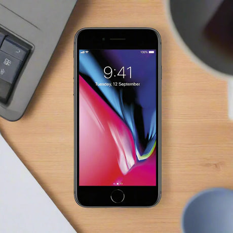 An Apple iPhone 8 64GB Space Grey Unlocked Smartphone (AU STOCK, Wty) by Apple rests on a wooden desk near a laptop keyboard, paper, and coffee cup, displaying the lock screen with the time 9:41 and date Tuesday, 12 September.