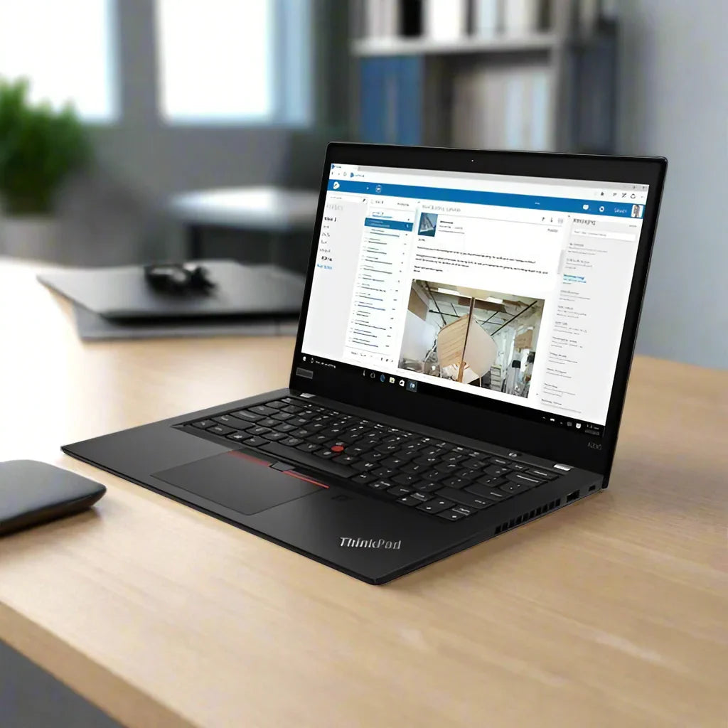 A black Lenovo ThinkPad X390 i5 8365U 1.6GHz 16GB 256GB SSD 13.3" Touch W11P laptop sits open on a wooden desk, displaying a website with a boat image; blurred shelves, a plant, and a closed laptop are in the background.