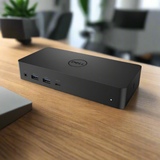A certified refurbished Dell D6000 USB 3.0 HDMI Display Ports Docking Station by Dell rests on a wooden desk with visible front ports, office furniture and potted plants blurred behind—ideal for versatile multi-monitor setups. Adapter included.