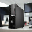 A certified refurbished Dell OptiPlex 7000 SFF i7-12700 2.1GHz 32GB 256GB desktop sits on a white office desk, with blurred monitors and equipment in the background.