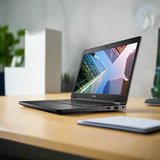 A black Certified Refurbished Dell Vostro 3578 i7 8550U laptop with 8GB RAM and 256GB SSD sits open on a wooden desk, showing skyscrapers on its 15.6 screen, with blurred office items, plants, and a closed notebook nearby.