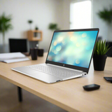 A certified refurbished HP 15S-EQ3XXX laptop (Ryzen 5 5625U, 16GB RAM, 512GB SSD, 15.6" W11P) sits on a wooden desk in a modern office, surrounded by a potted plant, smartphone, and supplies with shelves and plants behind.
