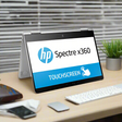 An HP Spectre x360 13-w031tu laptop with a slim aluminum body is set in tent mode on a desk, displaying “HP Spectre x360” and “TOUCHSCREEN” on its 13.3 screen. A keyboard, mouse, and plant are nearby.