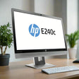 The HP EliteDisplay E240C 23.8 IPS monitor with webcam shows the HP logo and model on screen, a keyboard sits on the desk, and green potted plants appear in the background.