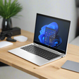 An HP EliteBook x360 830 G10 i5-1345U 16GB 256GB 13.3" Touch W11P laptop displays the Windows 11 start menu on a wooden desk, alongside a closed mouse, gray mouse pad, sunglasses, and a potted plant.
