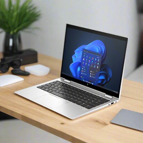 An HP EliteBook x360 830 G10 i5-1345U 16GB 256GB 13.3" Touch W11P laptop displays the Windows 11 start menu on a wooden desk, alongside a closed mouse, gray mouse pad, sunglasses, and a potted plant.