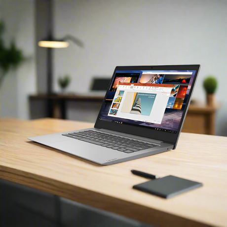 A slim Lenovo IdeaPad Slim 1-14AST-05 is open on a wooden desk with a stylus and closed notebook nearby; a blurred office scene with plants and a lamp is visible in the background.