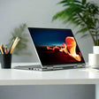A certified refurbished Lenovo ThinkPad X1 Yoga G6 i5 1135G7 16GB 256GB SSD 14 convertible laptop sits in tent mode on a white desk, displaying vibrant colors, with a potted plant and pencil cup in a modern workspace.