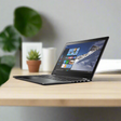 A slim black Lenovo Yoga 510-14ISK 14" touchscreen convertible laptop with Windows 10 Home is open on a light wooden desk, next to a small potted plant and a blurred large green leaf in the background.