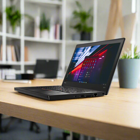 A refurbished black Lenovo ThinkPad X270 i5-7200U 4GB 500GB HDD 12.5 LTE W10P sits open on a wooden desk in a modern office, with blurred shelves, another laptop, and a potted plant nearby. The screen displays a colorful interface.