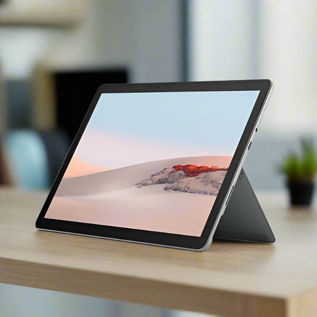 A Microsoft Surface Go 2 (4425Y, 1.7GHz, 8GB RAM, 128GB, 10.5 Touch) sits on a light wooden desk displaying sand dunes. Powered by Windows 11 Pro, it combines versatility and style amid a softly blurred background with a plant visible.