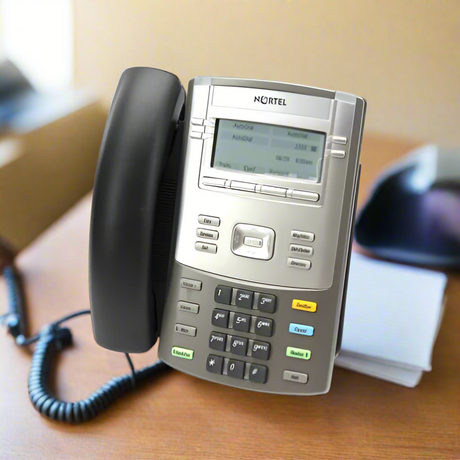 A Nortel IP Phone 1120E NTYS03, certified refurbished and sold without a power adapter, sits on a brown desk with its digital display, multiple buttons, notepaper stack, and a black object that may be a mouse. 3-month warranty included.
