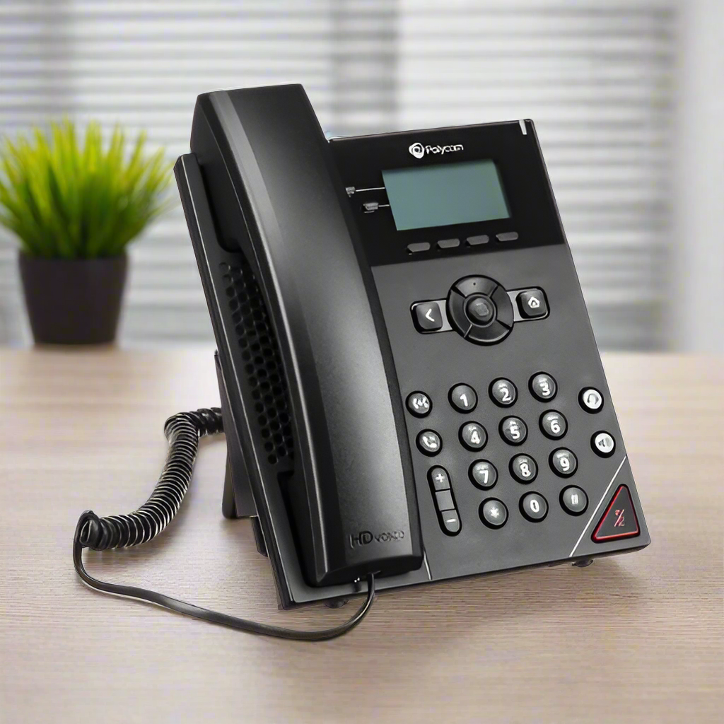 A black Polycom VVX 150 2-Line Business VoIP IP Desk Phone with corded handset sits on a wooden desk, with a blurred green potted plant in the background.