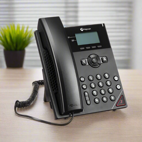 A black Polycom VVX 150 2-Line Business VoIP IP Desk Phone with corded handset sits on a wooden desk, with a blurred green potted plant in the background.