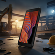 A Samsung Galaxy XCover 5 64GB 4GB Rugged Smartphone by Samsung stands upright on a construction site table with tools and a hard hat, while excavators and buildings appear in the background under bright sunset work lights.