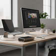 A bright office workspace features a Targus DOCK120 USB 3.0 Universal Docking Station, a desktop, an open laptop on a stand, a smartphone, and potted plants on a light wood desk near large windows.