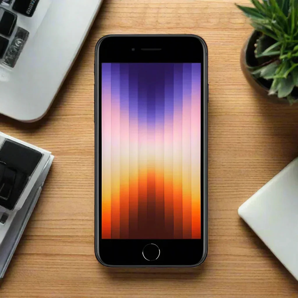 An Apple iPhone SE 2022 3rd Gen 64GB Midnight (LOCKED) with a striped gradient wallpaper rests on a wooden desk beside a keyboard, plant, and closed laptop.