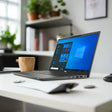 A Dell Latitude 7420 Laptop 14" (i5-1135G7, 8GB, 256GB SSD, Win 11 Pro) by Dell sits on a white desk with a mug, notebook, and pen in a bright modern office with shelves and plants in the background.