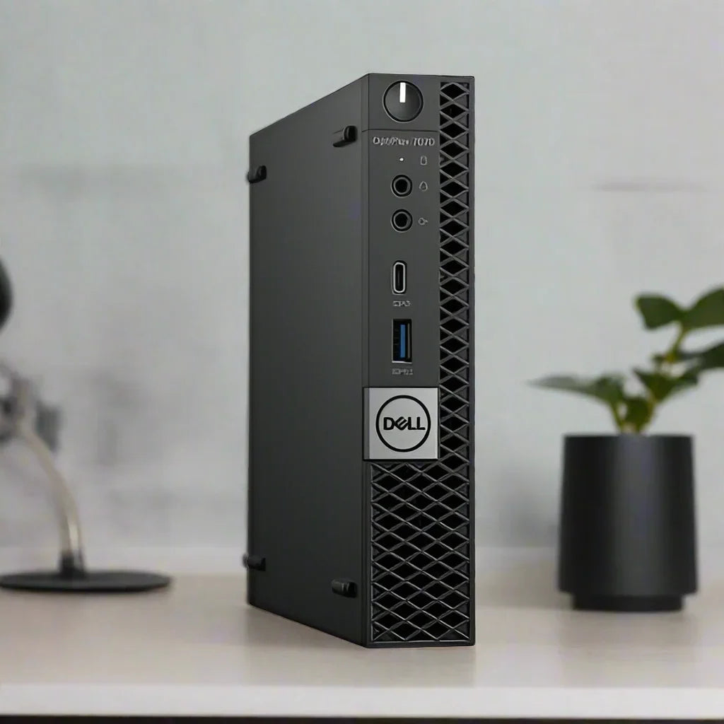 A Dell OptiPlex 7070 Micro i5 9500T desktop stands vertically on a desk, with a potted plant and blurred lamp in the background. Its front panel shows several ports and features the Dell logo for this compact business PC.