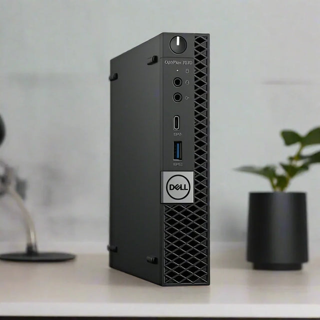 A Dell OptiPlex 7070 Micro i5 9500T desktop stands vertically on a desk, with a potted plant and blurred lamp in the background. Its front panel shows several ports and features the Dell logo for this compact business PC.
