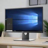 A refurbished Dell P2417H IPS 24 1920x1080 monitor sits on a light desk, showing the Windows 10 desktop with the Cortana search bar. The background is slightly blurred, revealing a wall and some green plants. No stand included.