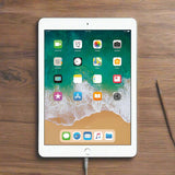 An Apple iPad 6th Gen 9.7 128GB WIFI Silver tablet from Apple is lying on a wooden surface, charging, with app icons on its home screen and a metal tool resting beside it.