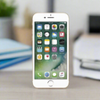 An Apple iPhone 7 256GB Silver Unlocked Smartphone (A-Grade, 6-month warranty) stands upright on a desk with its home screen visible; the background shows blurred stacked books and a green plant.