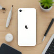A refurbished white Apple iPhone SE 2020 (128GB, Unlocked, AU Stock) with a black logo lies face down on a wooden desk beside a pen, screwdriver, smart device, potted plant, and keyboard. Powered by the A13 Bionic chip.