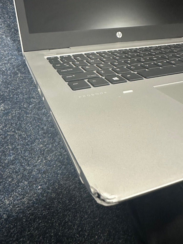 A silver HP ProBook 640 G5 i5 8365U laptop (16GB RAM, 256GB, WWAN, Windows 11 Pro) with a front left corner dent and exposed material is visibly damaged and placed on a dark blue carpeted floor. Certified refurbished by HP.