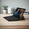 A refurbished Microsoft Surface Pro 4 1724 (i5 6300U, 8GB RAM, 256GB SSD, 12 Touch, Windows 10 Pro) with keyboard sits on a wooden desk. Its display shows scenic wallpaper; a potted plant is blurred in the background.