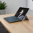A refurbished Microsoft Surface Pro 5 i7 7660U 2.5GHz, 8GB RAM, 256GB, 12.3 W10P by Microsoft with a keyboard cover and built-in stand rests on a wooden table; the lock screen shows a crane bird with a blurred green plant behind it.