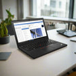 A black Lenovo ThinkPad X260 i5 6300U 2.4GHz 8GB 256GB SSD W10P 12.5" laptop is open on a white desk with a potted plant, smartphone, notebook, and pen, in a bright office with large windows.